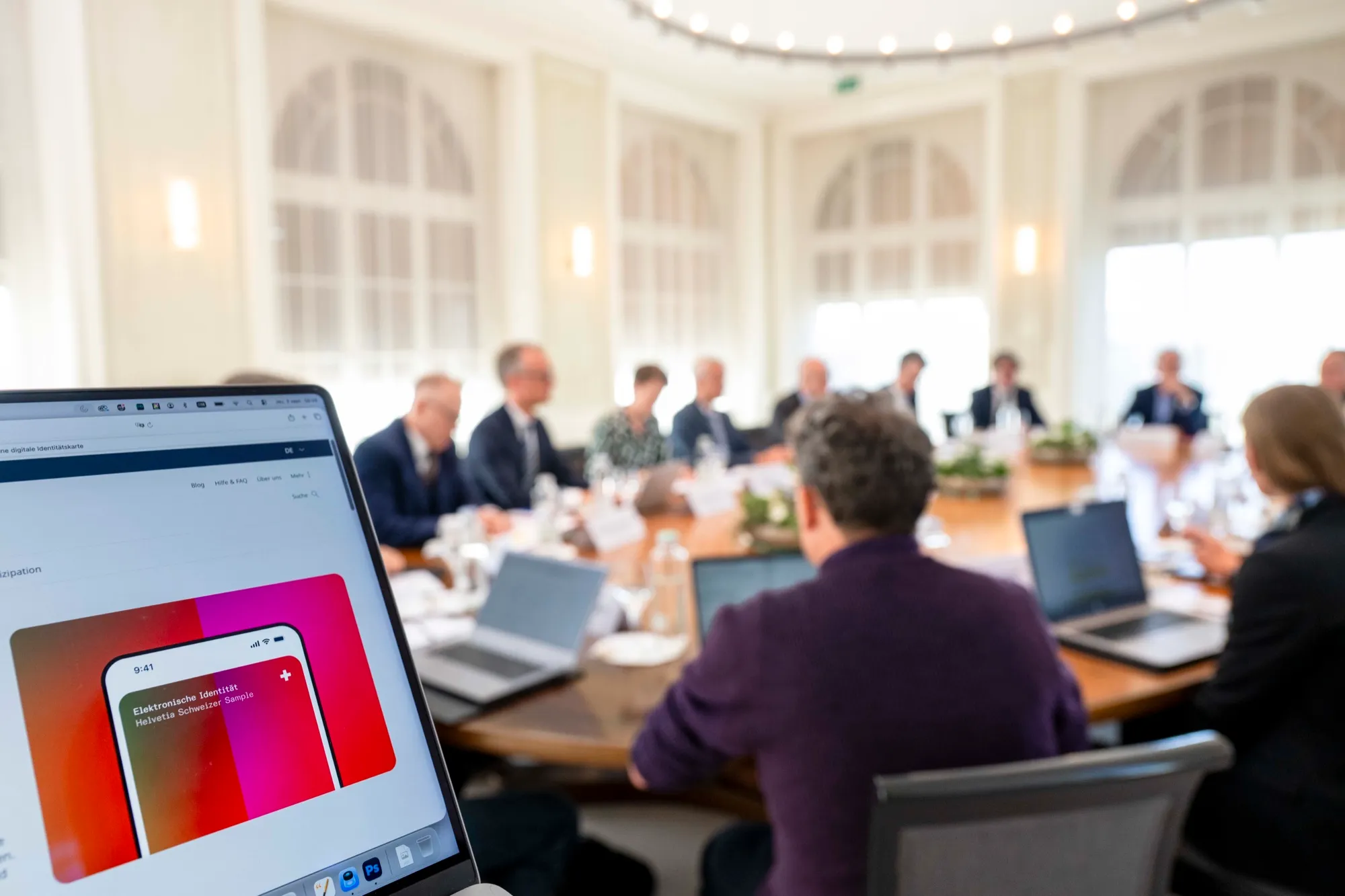 Laptop mit e-ID-Grafik im Vordergrund, dahinter Teilnehmende des Beiratstreffens an einem langen Tisch in einem hellen Saal