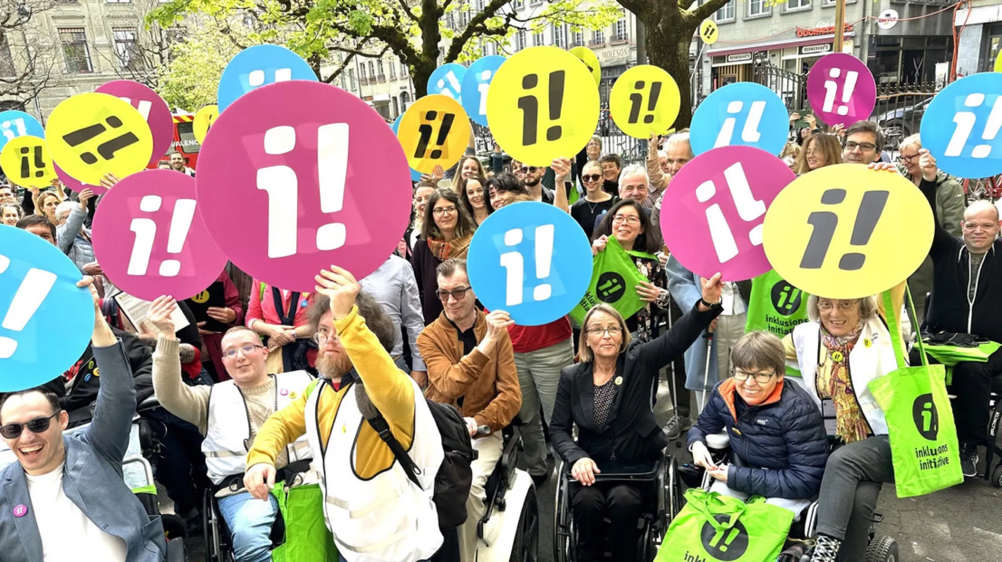 Grosse Menschenmenge, viele im Rollstuhl, hält bunte runde Schilder mit dem Logo der Inklusions-Initiative hoch