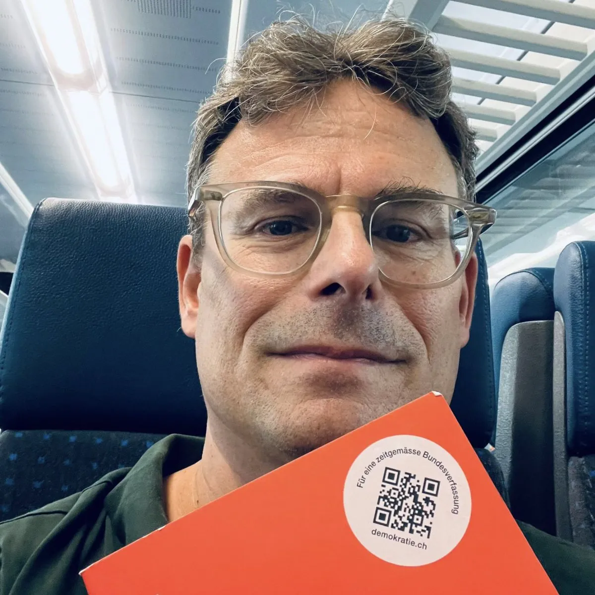 Daniel Graf im Zug mit einem orangen Heft «Für eine zeitgemässe Bundesverfassung»